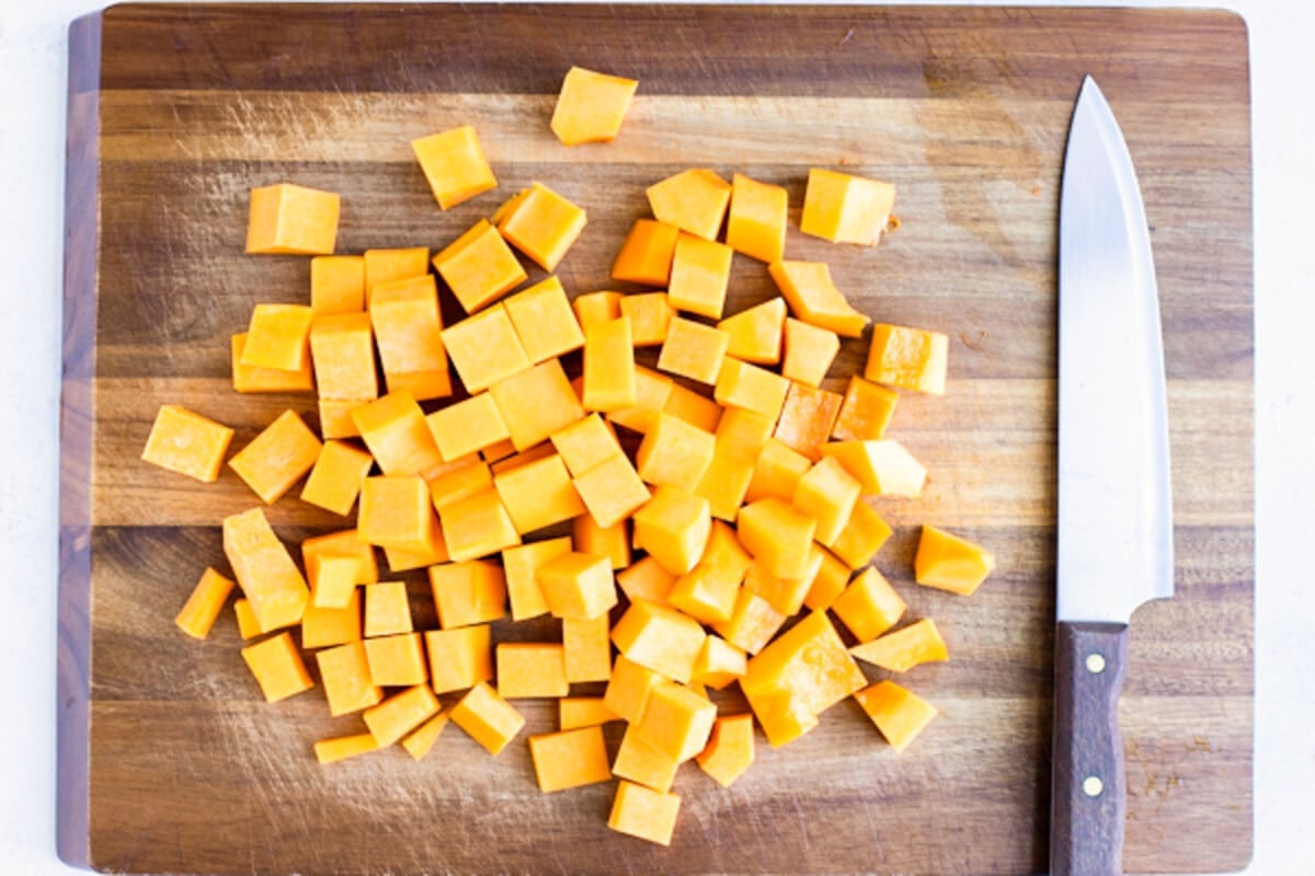 Cubed butternut squash is on a cutting board with a knife for a roasted recipe.
