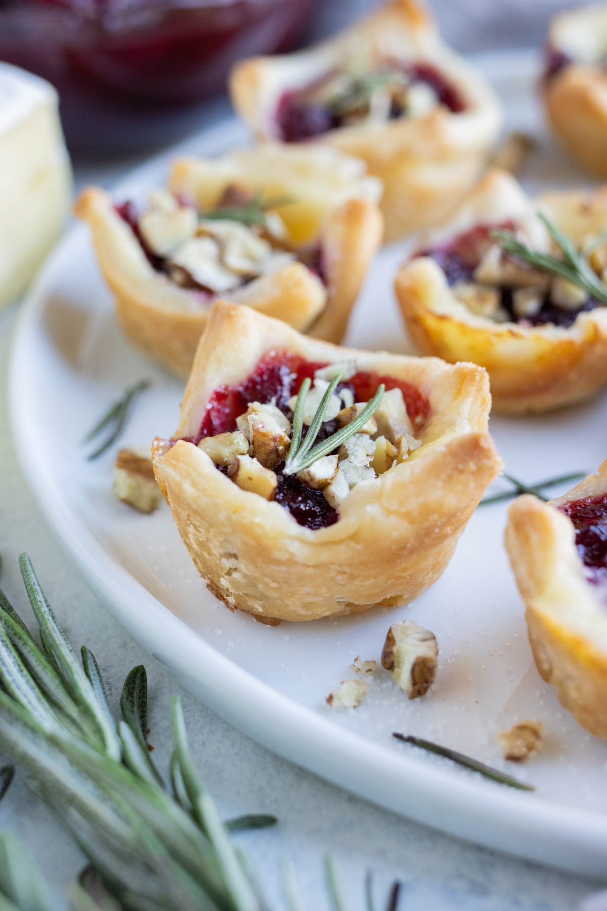 A plate of brie bites with cranberry is set on the counter near fresh rosemary.