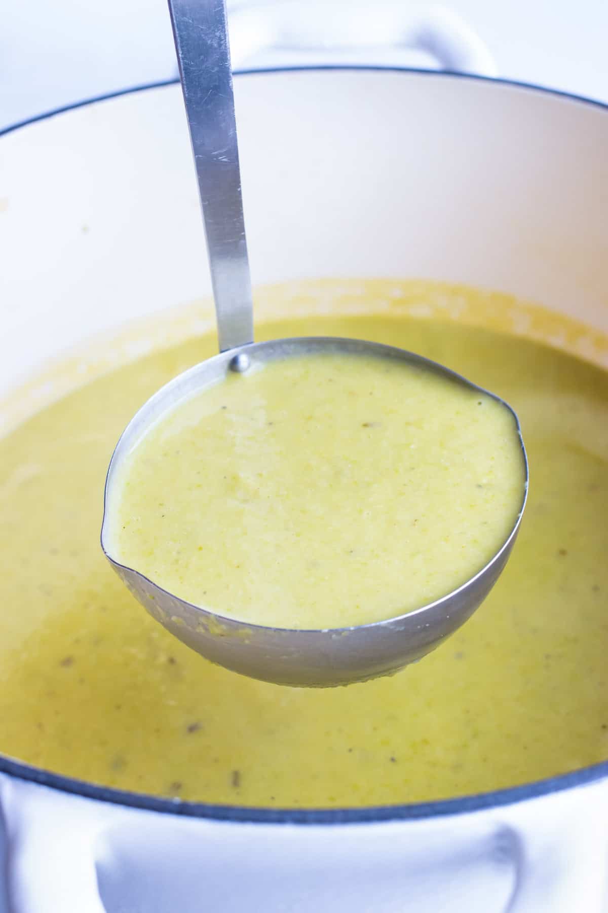 A ladle scooping out a serving of vegan asparagus soup from a white pot.