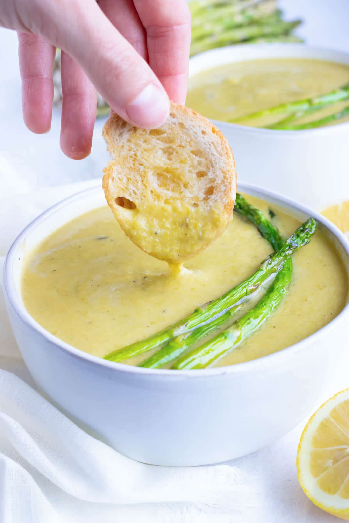 A toasted baguette being dipped into a bowl full of creamy asparagus soup.