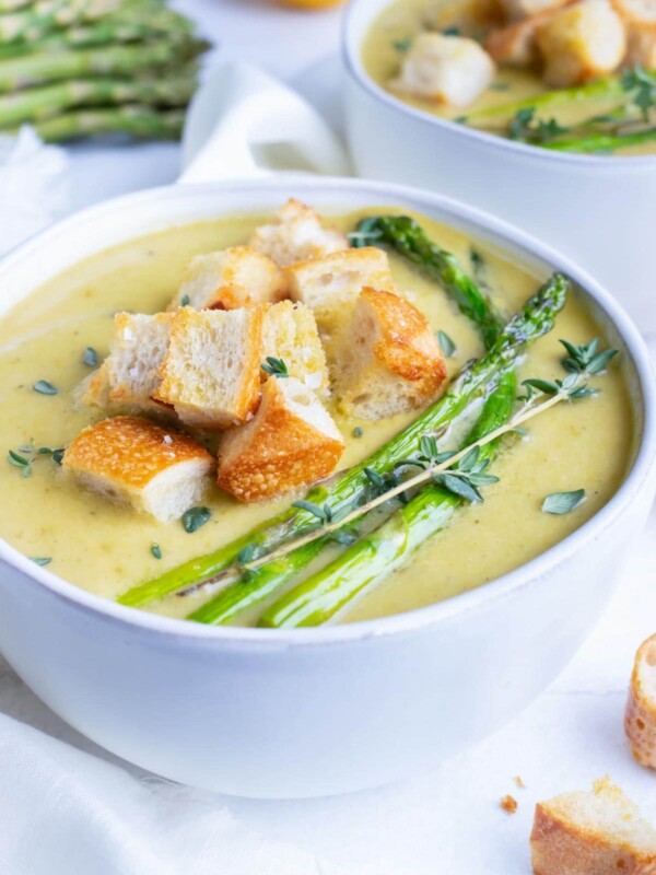 A white serving bowl full of a creamy asparagus soup recipe with roasted asparagus spears and homemade croutons.