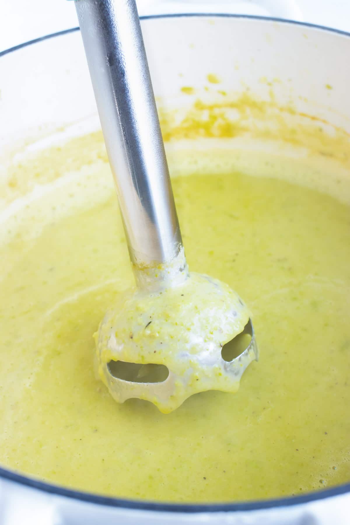 An immersion blender pureeing a batch of vegetable soup in a white Dutch oven.