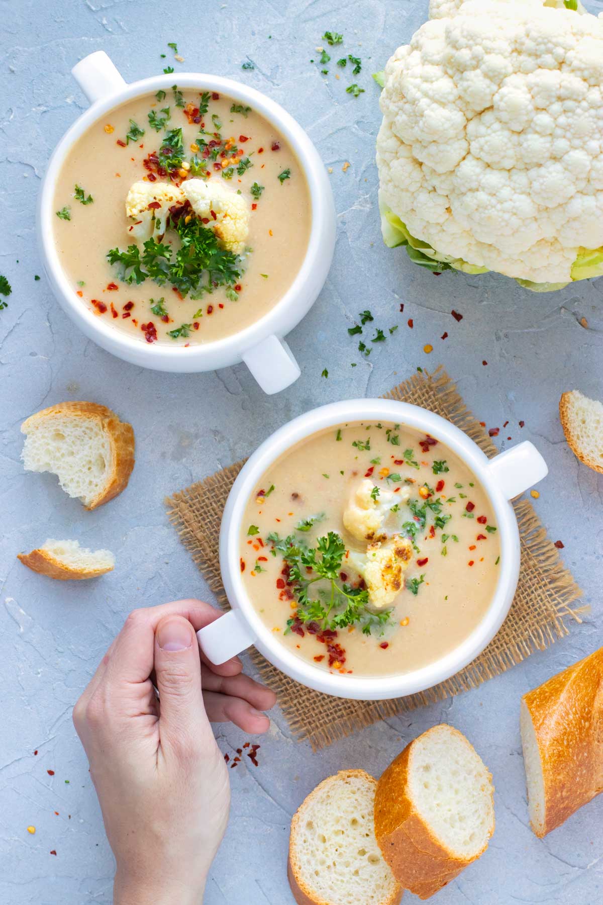 Two white bowls full of a healthy cauliflower soup recipe next to a head of cauliflower and served with a baguette.