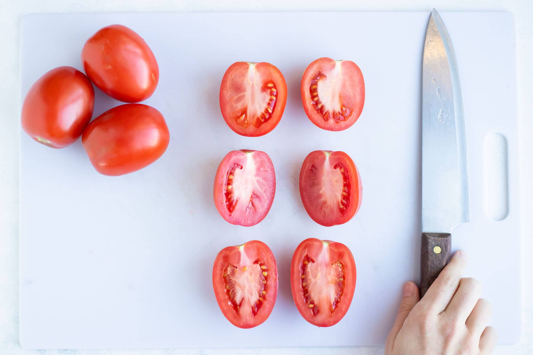 Halved tomatoes on a white cutting board next to a hand holding a knife.