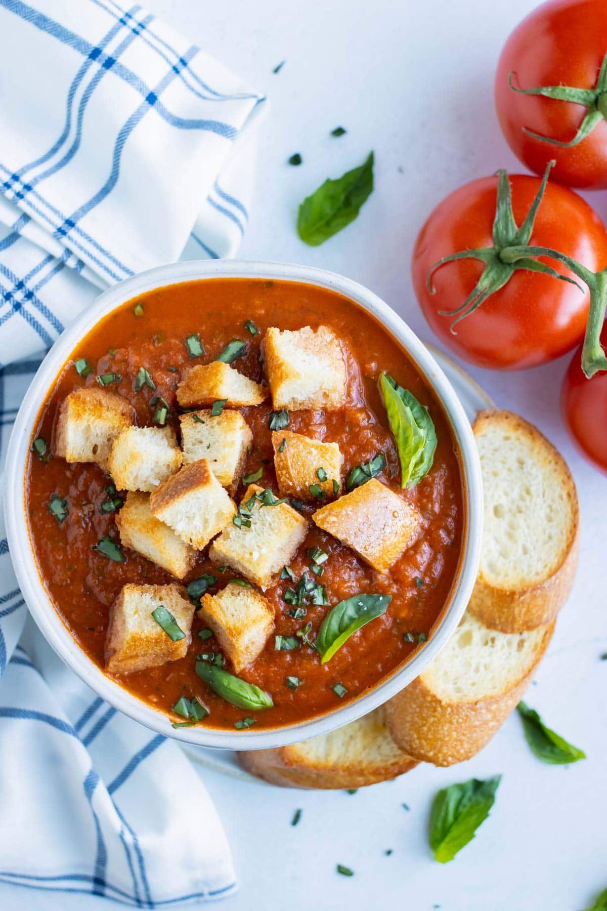 Vegan roasted tomato basil soup in a white bowl with toasted baguettes and fresh tomatoes next to it.