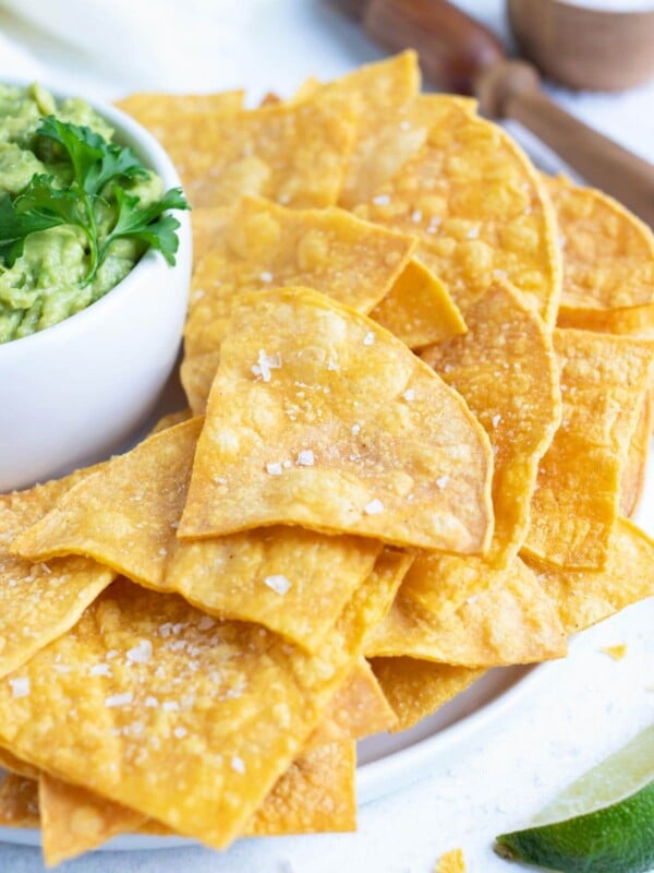 Homemade tortilla chips on a white plate next to a bowl of guacamole.