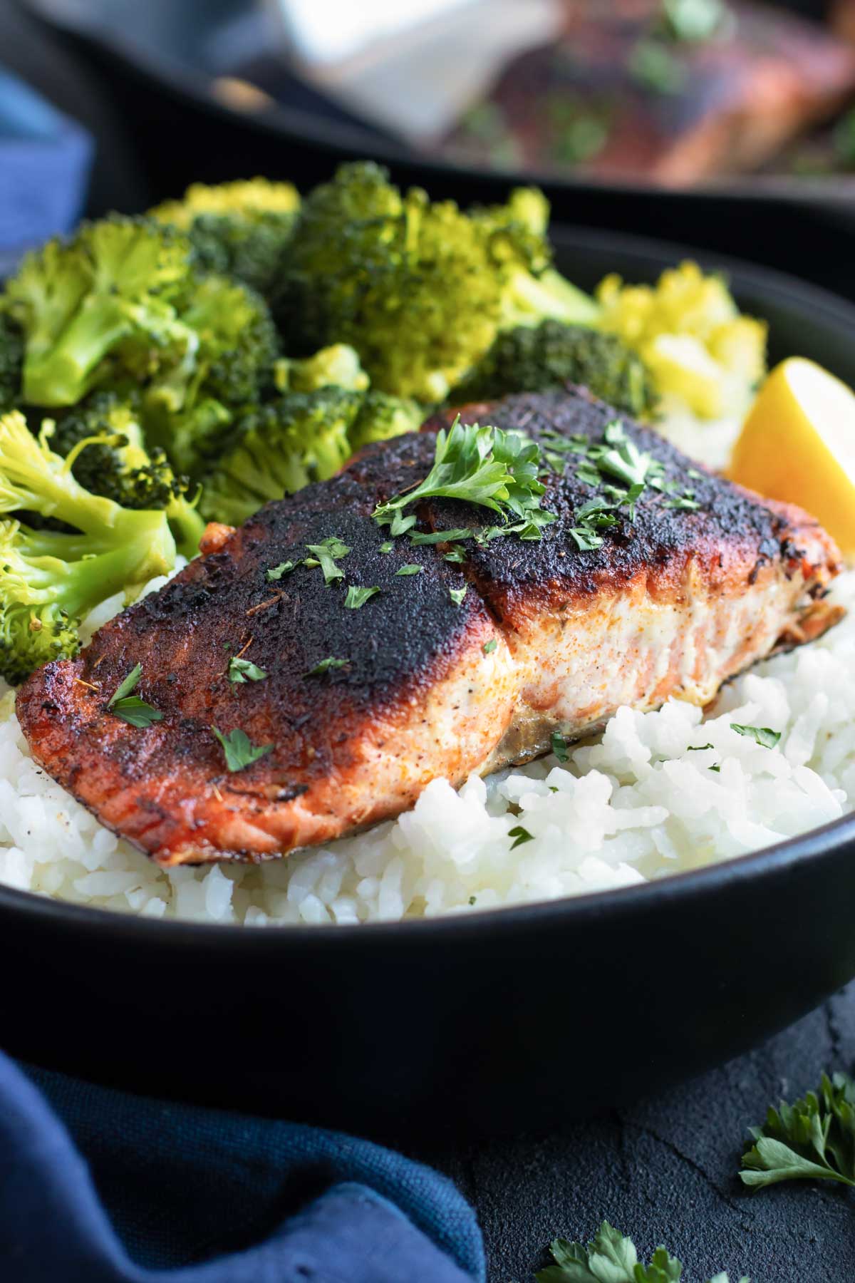 Baked salmon with blackened seasoning over a bed of rice with steamed broccoli in a black bowl.