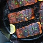 Cajun blackened salmon fillets in a cast-iron skillet with lemon wedges.
