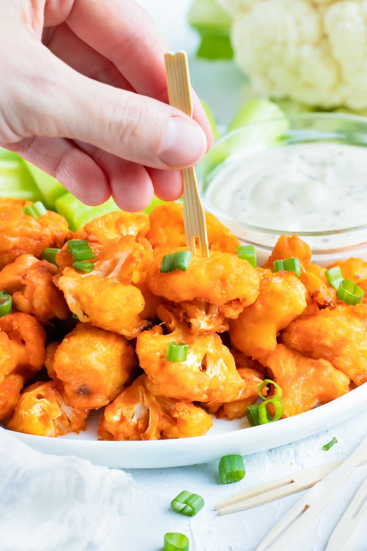 A wooden toothpick that is picking up a buffalo cauliflower from a white plate with ranch dressing in the background.