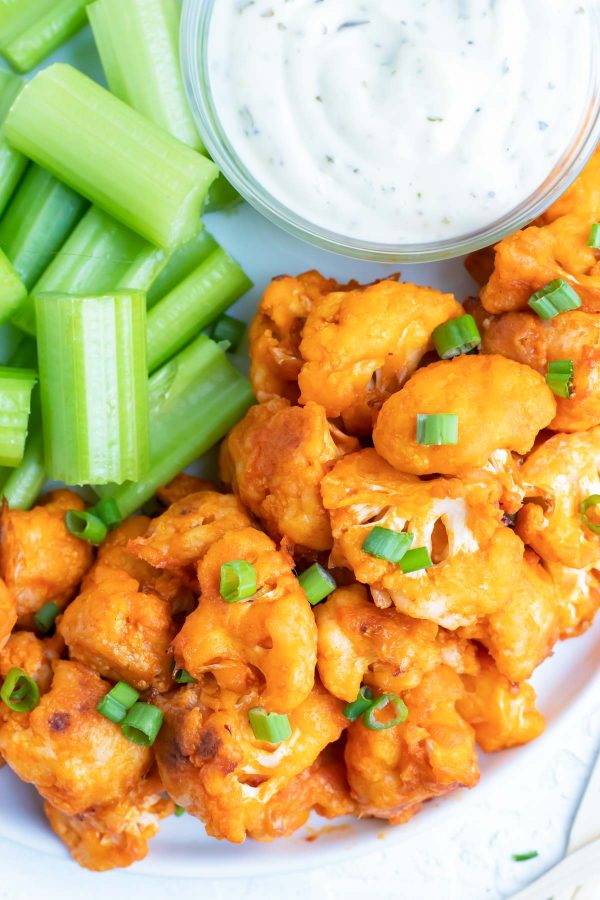 A white plate full of buffalo cauliflower bites, celery sticks, and a bowl full of ranch dressing for a Super Bowl appetizer.