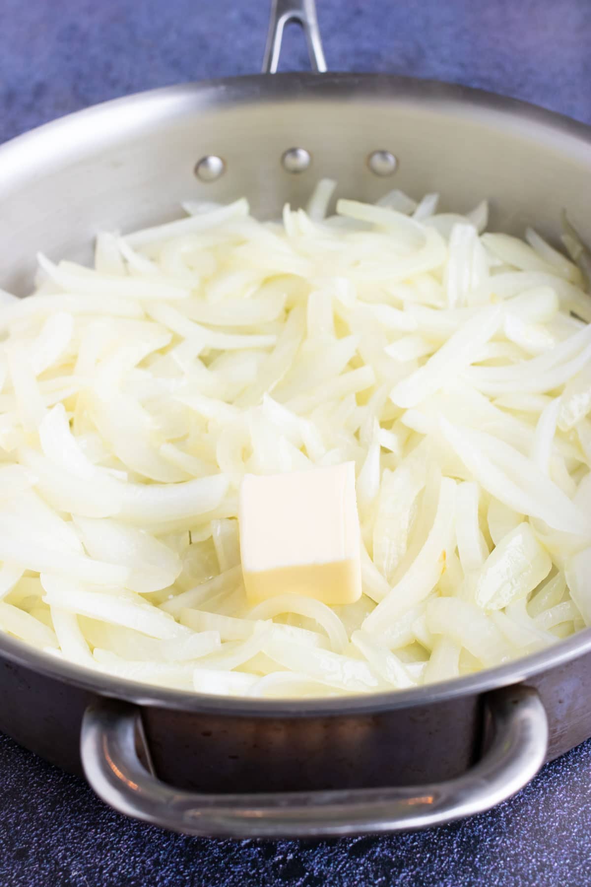 An image showing butter being added to partially cooked onions in a skillet.