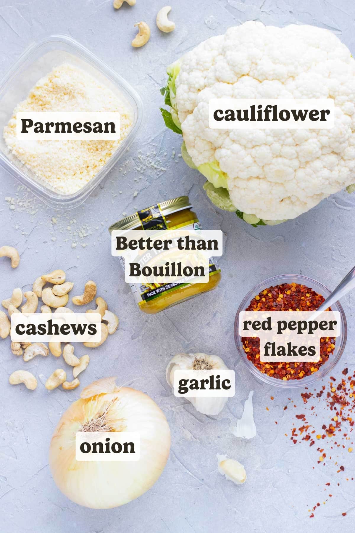 A head of cauliflower, Parmesan cheese, cashews, garlic, red pepper flakes, and an onion on a table as the ingredients for a cauliflower soup recipe.