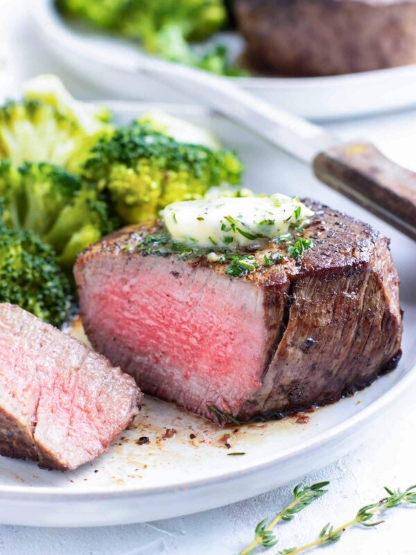 A filet mignon steak that has been cooked to medium-rare on a white plate with a knife and broccoli.