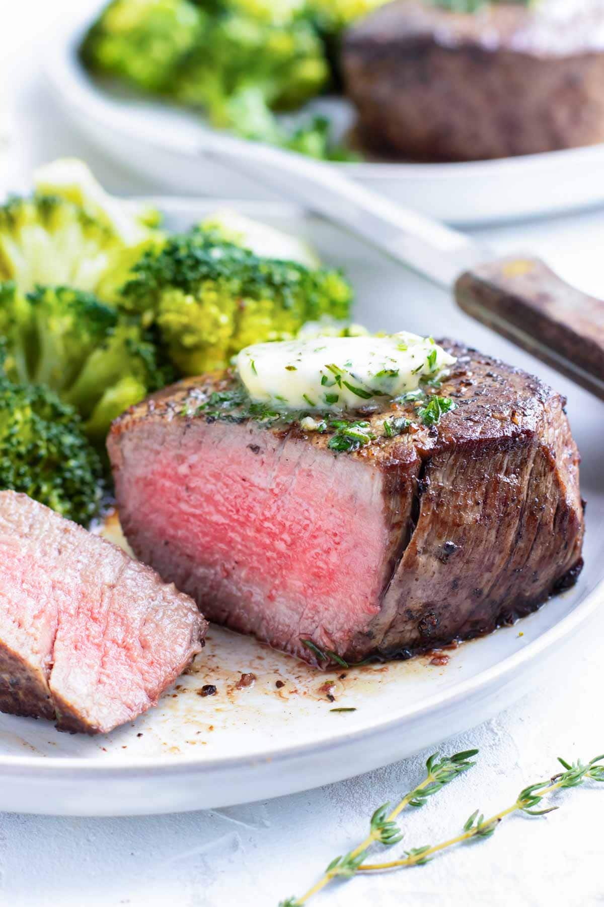 A filet mignon steak that has been cooked to medium-rare on a white plate with a knife and broccoli.