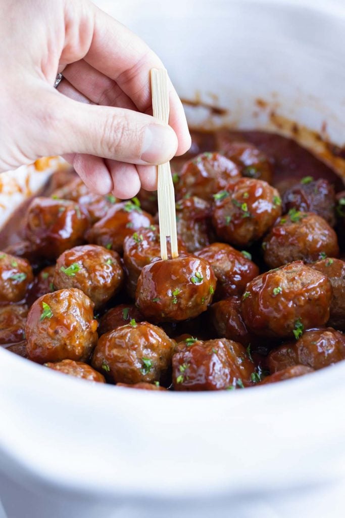 A hand is used to pick up a meatball with a toothpick.