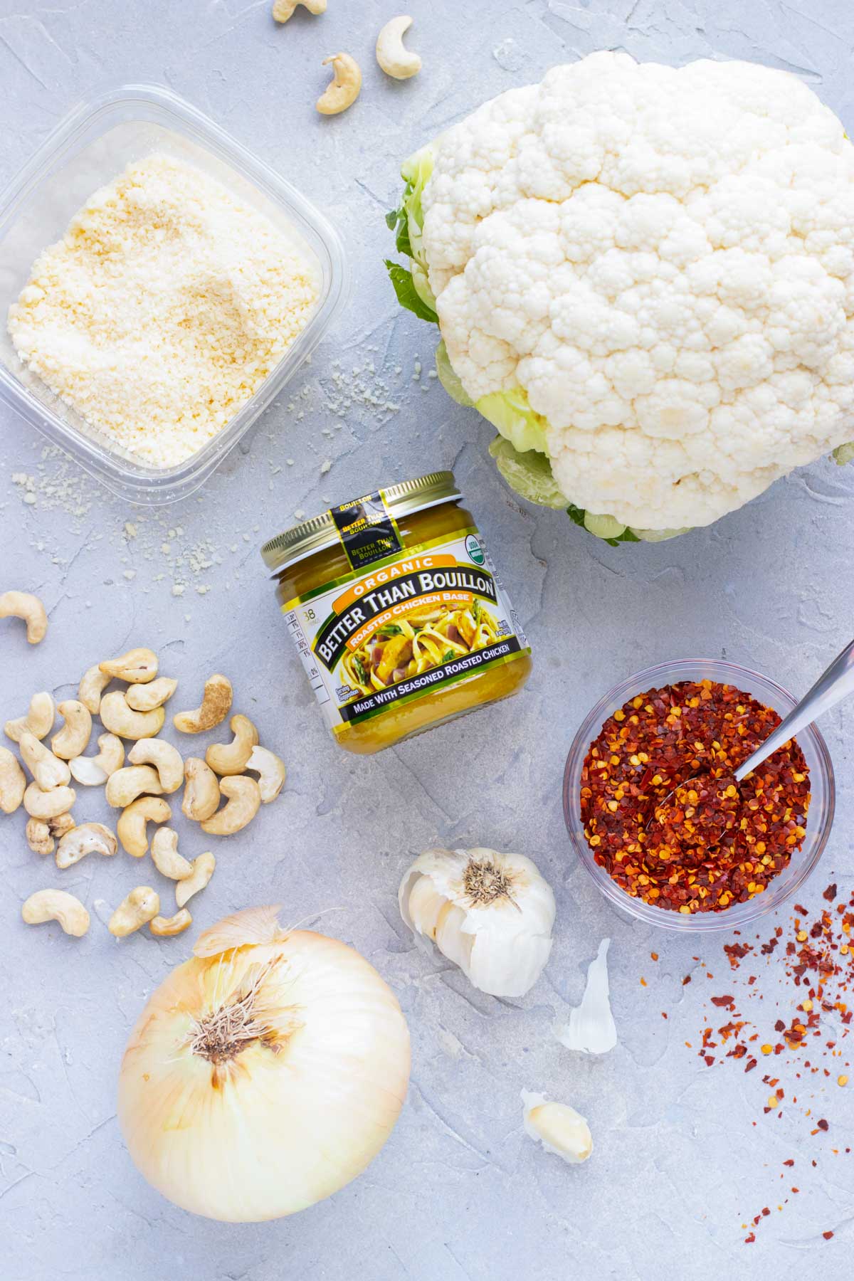 A head of cauliflower, Parmesan cheese, cashews, garlic, red pepper flakes, and an onion on a table as the ingredients for a cauliflower soup recipe.