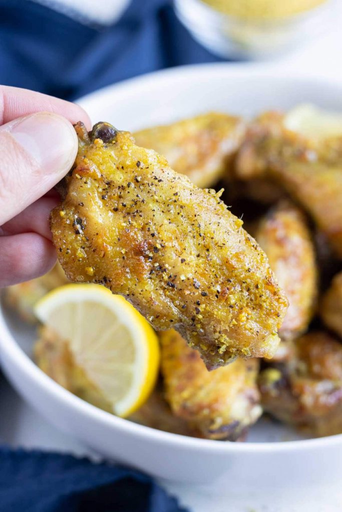 Lemon pepper chicken wings baked in the oven are picked up by a hand from a bowl.