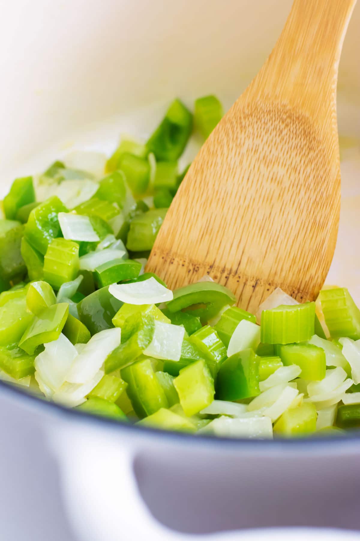 A wooden spatula sautés celery, bell pepper, and onion in a pot.