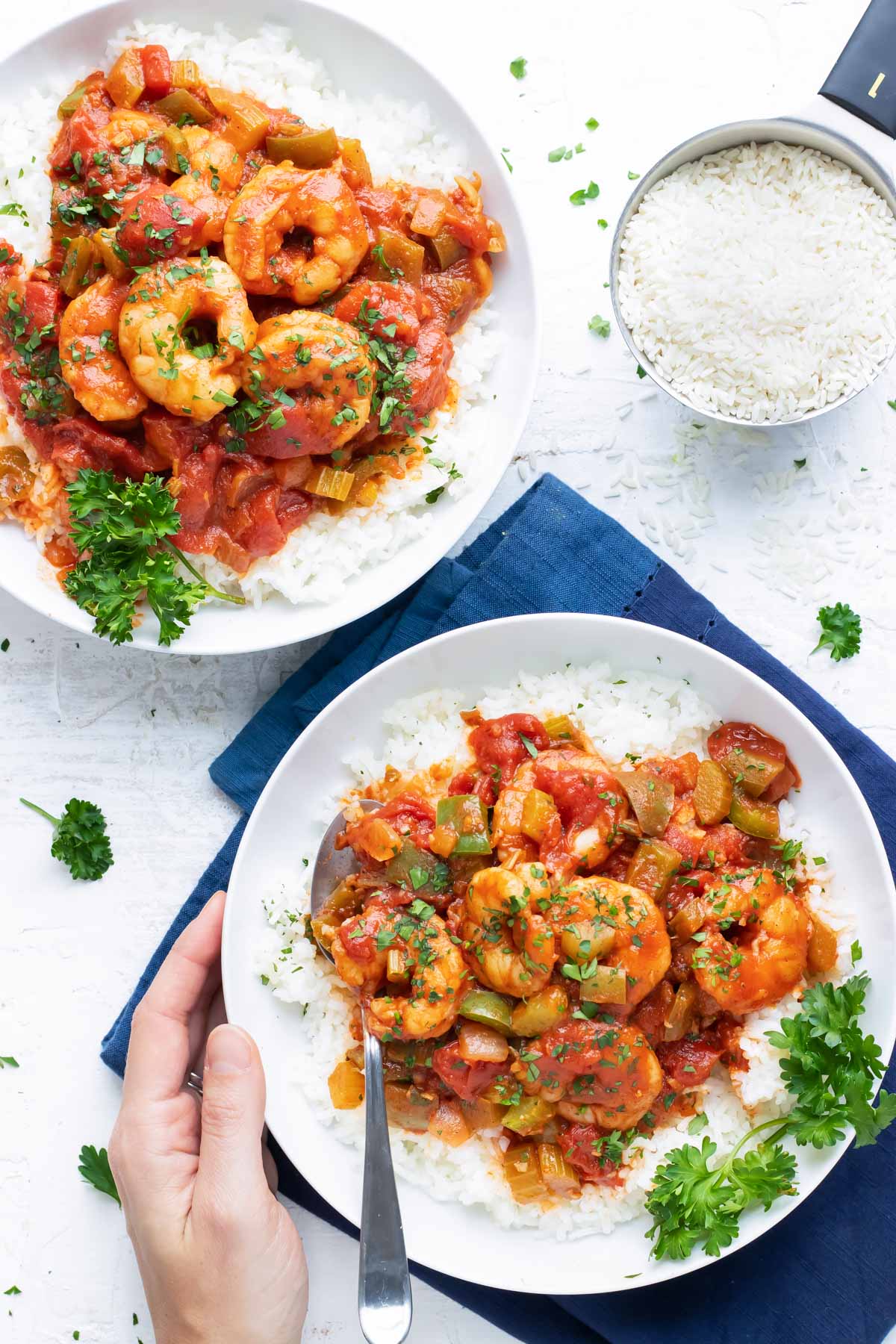 Two plates of shrimp creole and white rice being served, beside a measuring cup of uncooked rice.