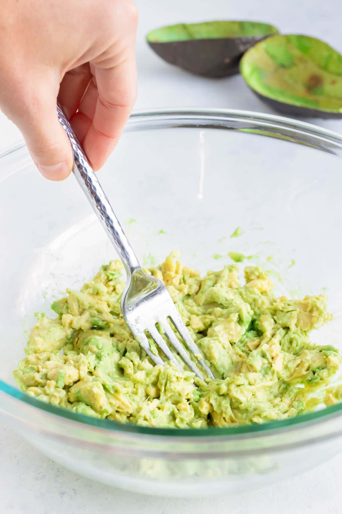 Ripe avocado is smashed by a fork in a glass bowl.