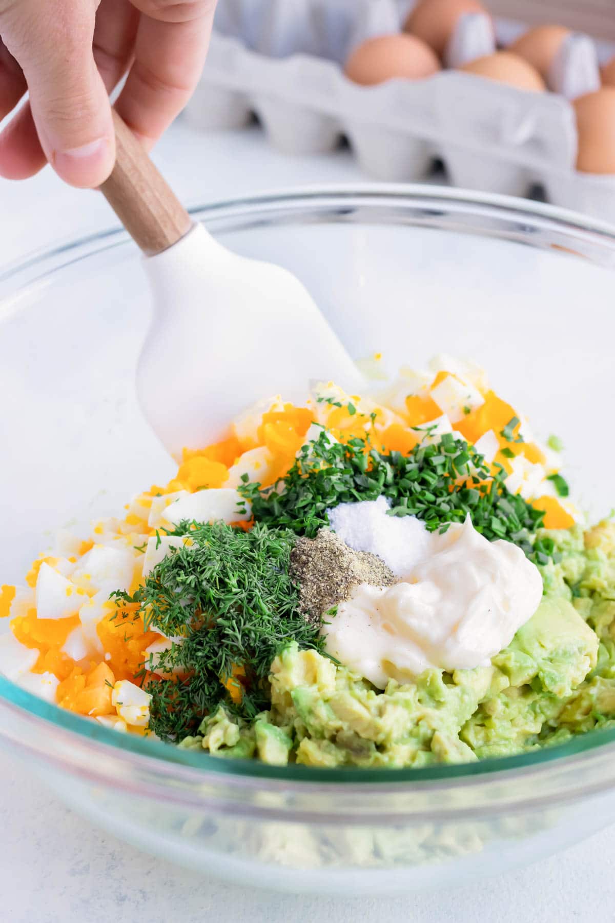 Fresh herbs, eggs, avocado, and mayonnaise are mixed together in a bowl.