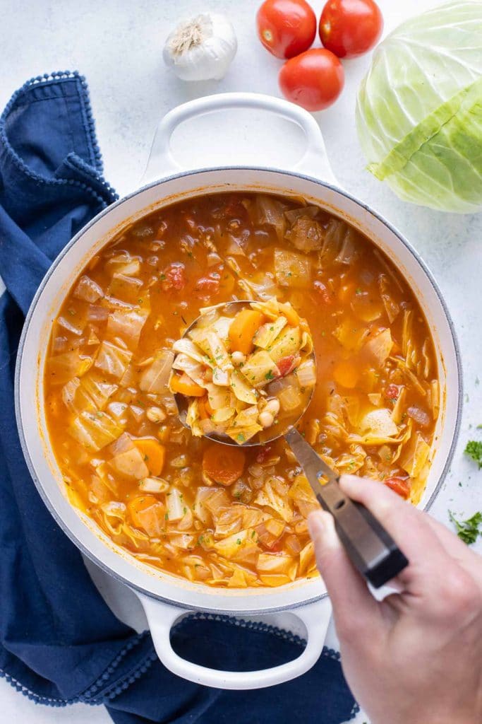 A ladle rests in a Dutch oven of homemade cabbage soup with white beans and carrots.