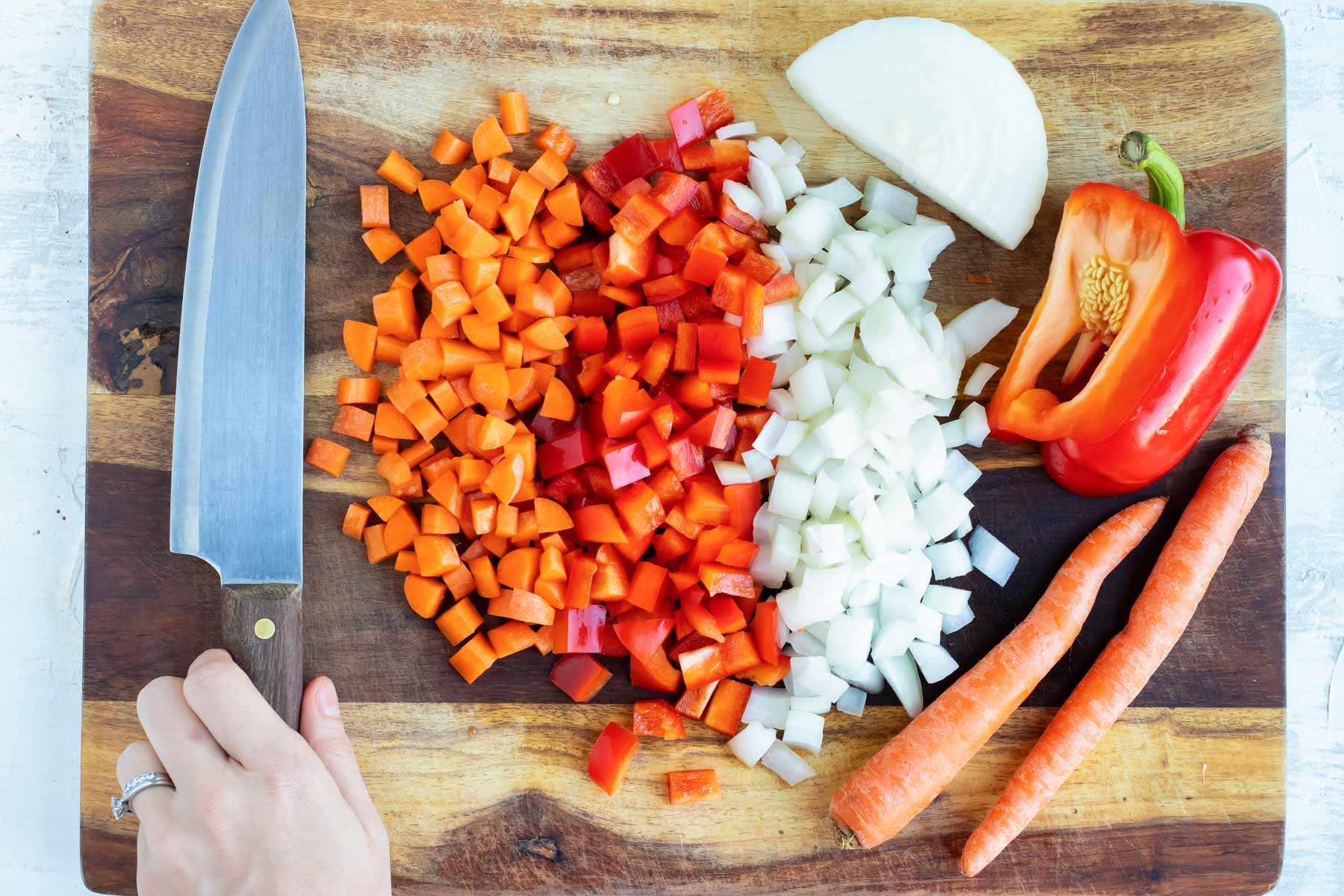 Diced carrots, red bell pepper, and onion sit on a wooden cutting board beside a knife.