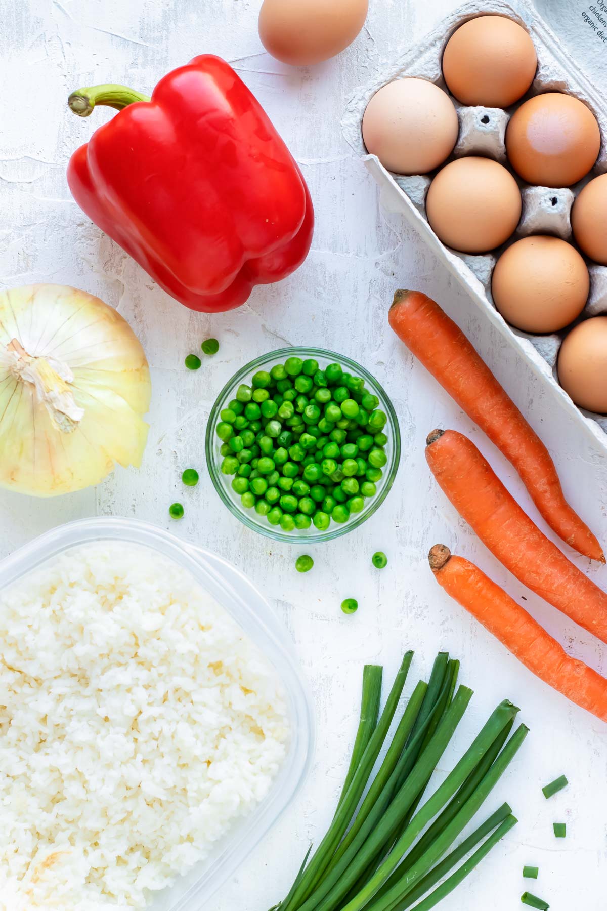 Eggs, rice, red bell pepper, onion, carrots, and green onion arranged on a white countertop.
