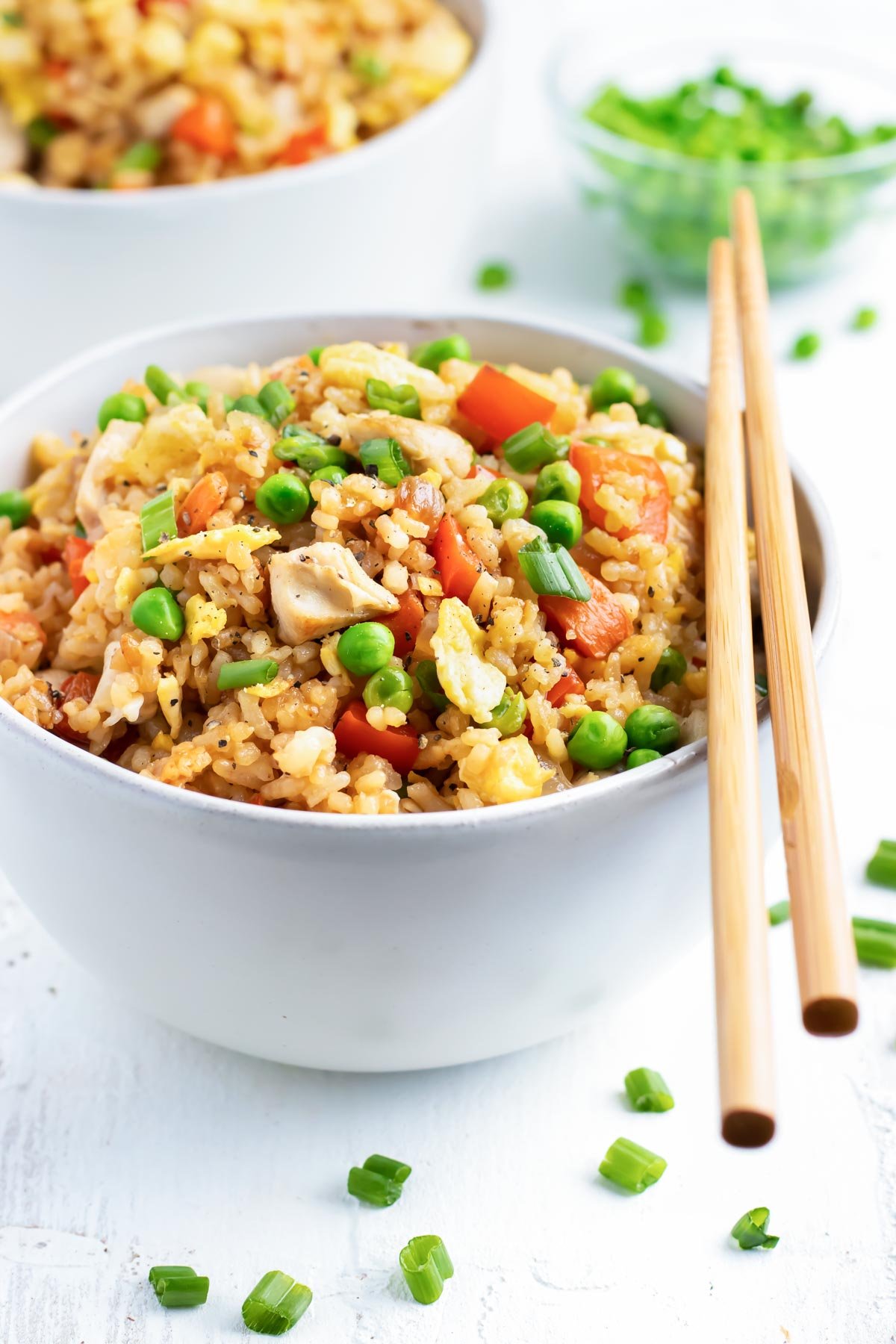 A white bowl full of a healthy fried rice recipe with carrots and peas next to green onions.