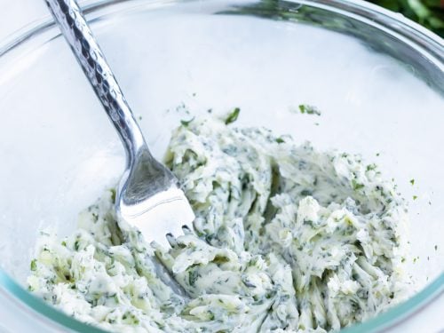 A fork is used to combine garlic herb butter ingredients together in a bowl.