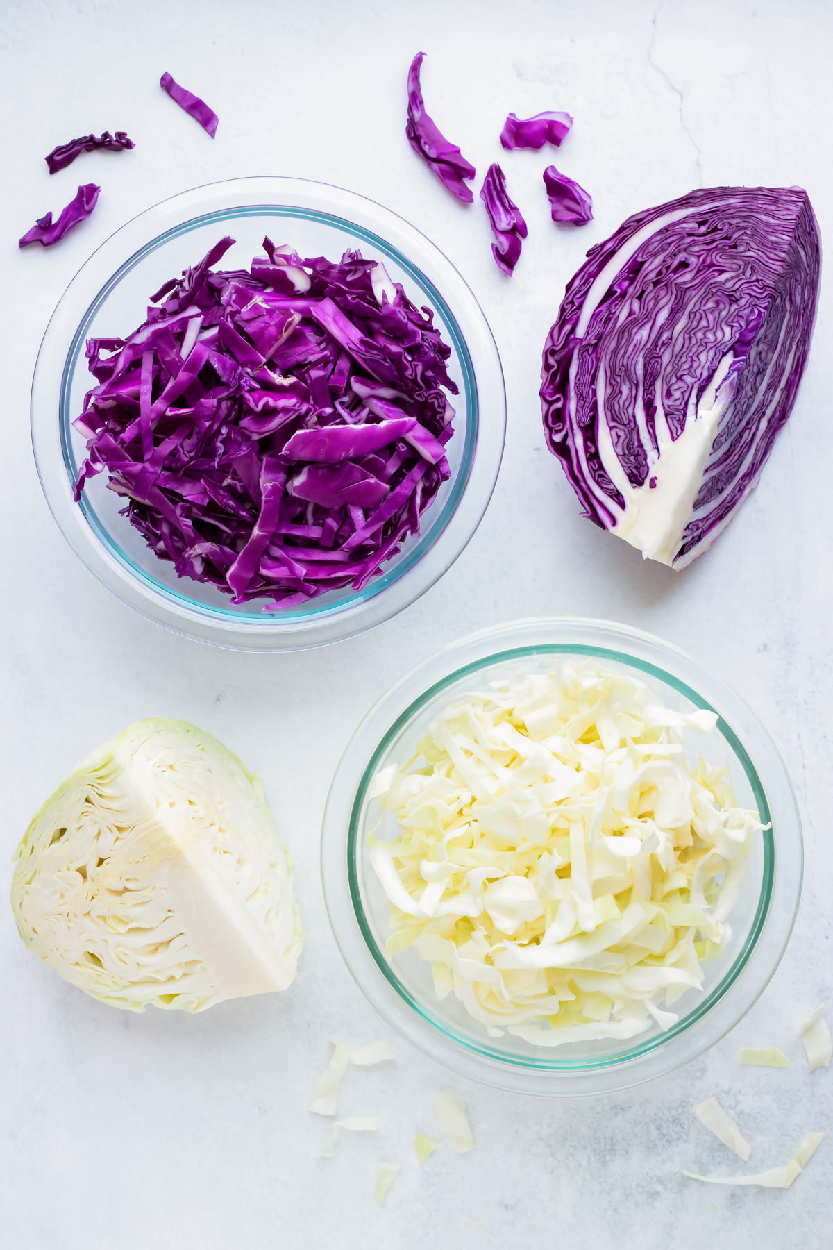 A bowl of shredded green cabbage beside a green cabbage wedge, and a bowl of shredded red cabbage beside a red cabbage wedge.