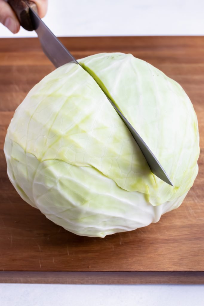 A head of cabbage with the stem side down on a cutting board being cut in half lengthwise.