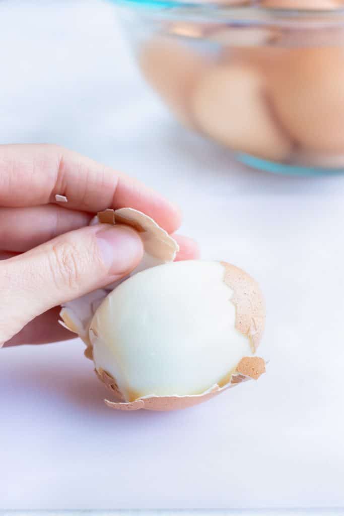 A hand peeling the shell from a hard-boiled egg for potato salad with eggs.
