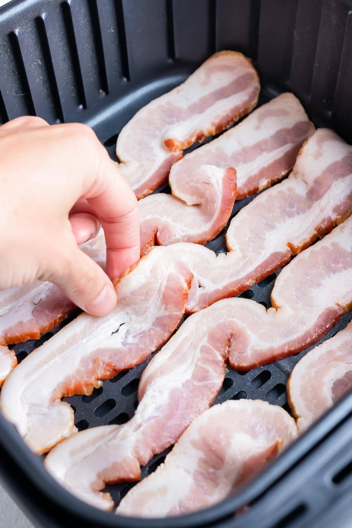 A hand positions bacon slices in a single layer in an air fryer basket so none are overlapping.