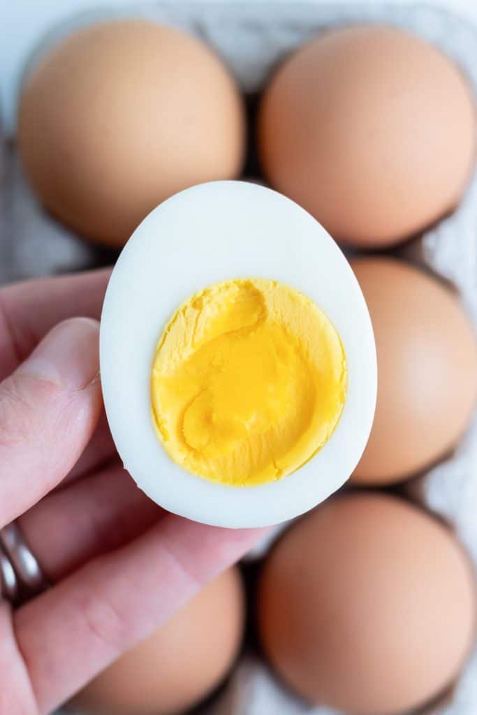 A hard-boiled egg is held by a hand with more eggs behind it.