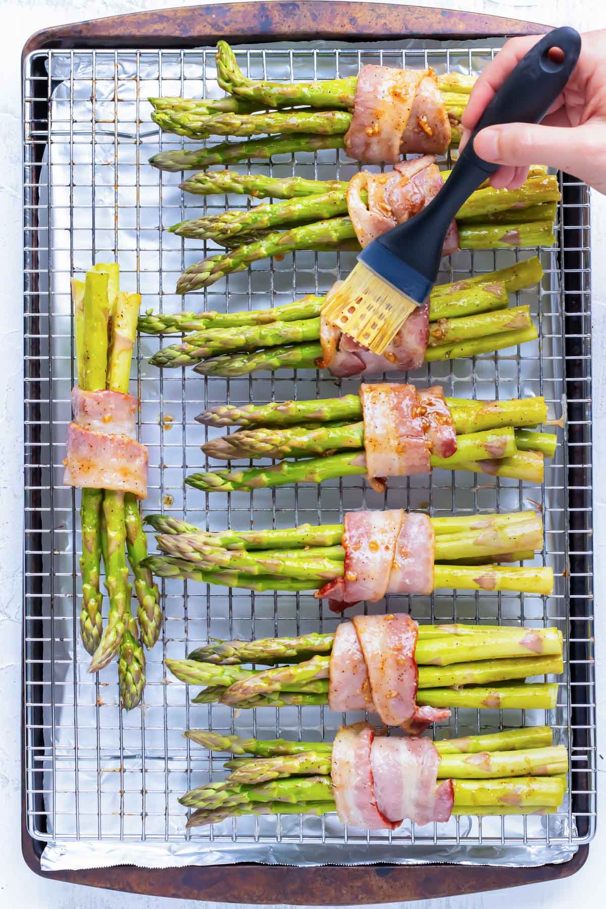 Garlic and butter sauce being brushed over bacon wrapped asparagus on a wire rack and baking sheet.