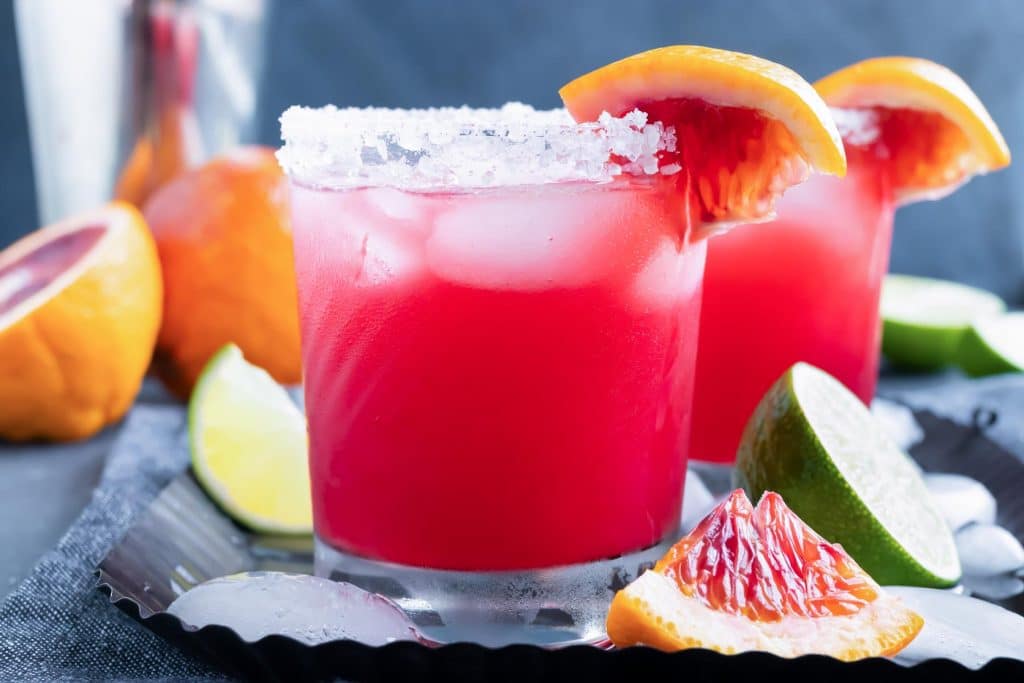 A homemade margarita in a clear glass with blood oranges in the background.