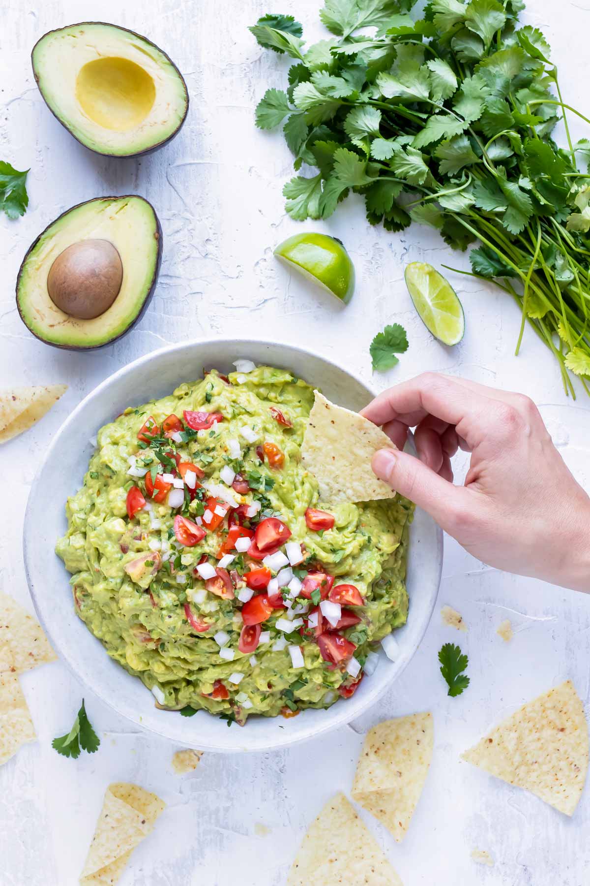 A hand dipping a corn tortilla chip into a big bowl full of a simple guacamole with tomatoes and onions.