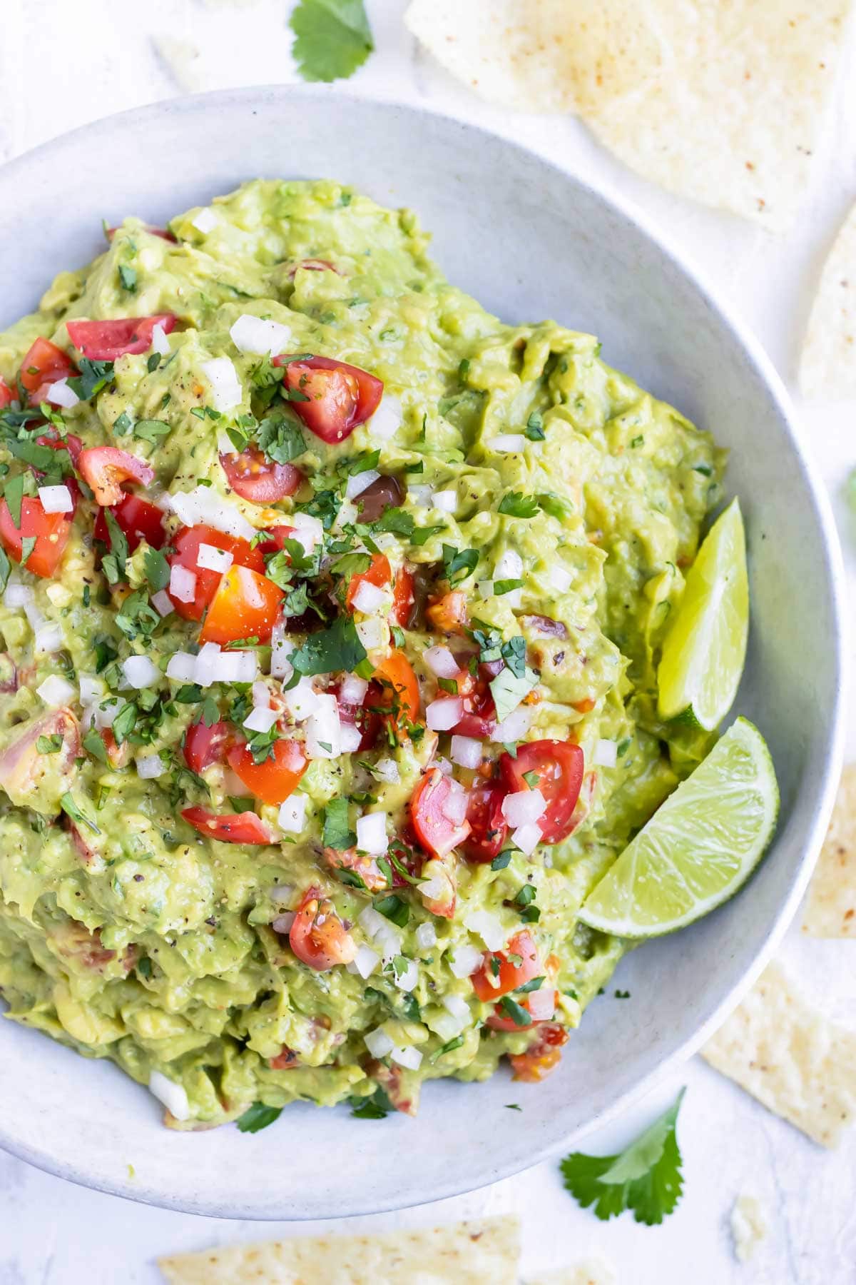 A white bowl full of a simple guacamole recipe with limes, tomatoes, and onions to garnish.