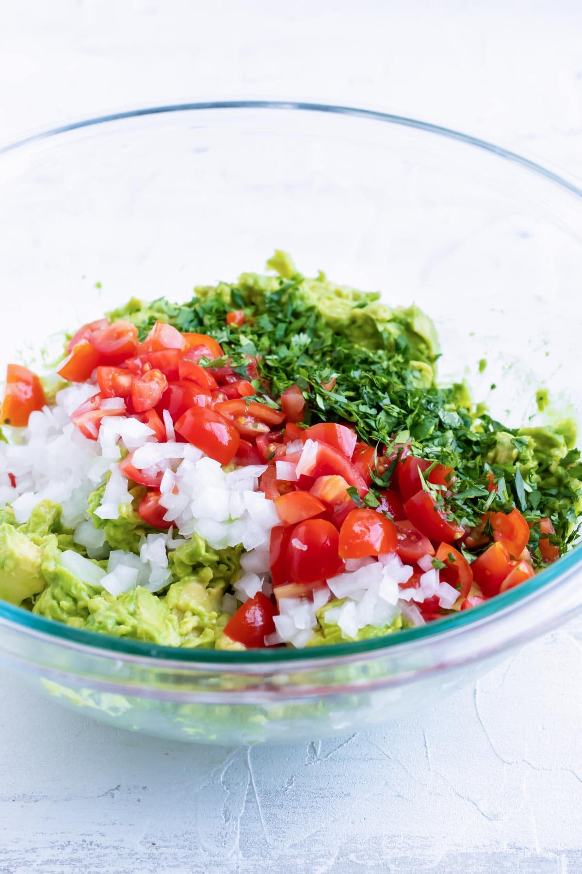 Combining onions, tomatoes, and cilantro to mashed avocados in a clear bowl.