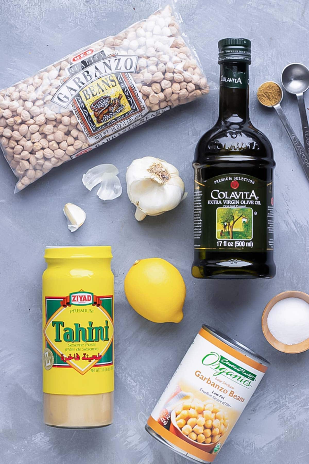 Hummus ingredients (garbanzo beans, olive oil, garlic, lemon, tahini, and salt) on a grey table.