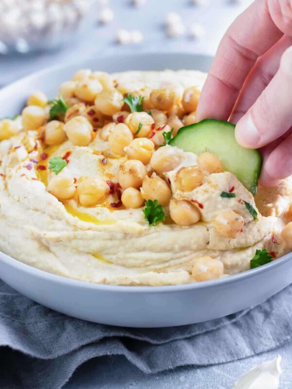 A cucumber slice being dipped into a party bowl full of a healthy authentic hummus.