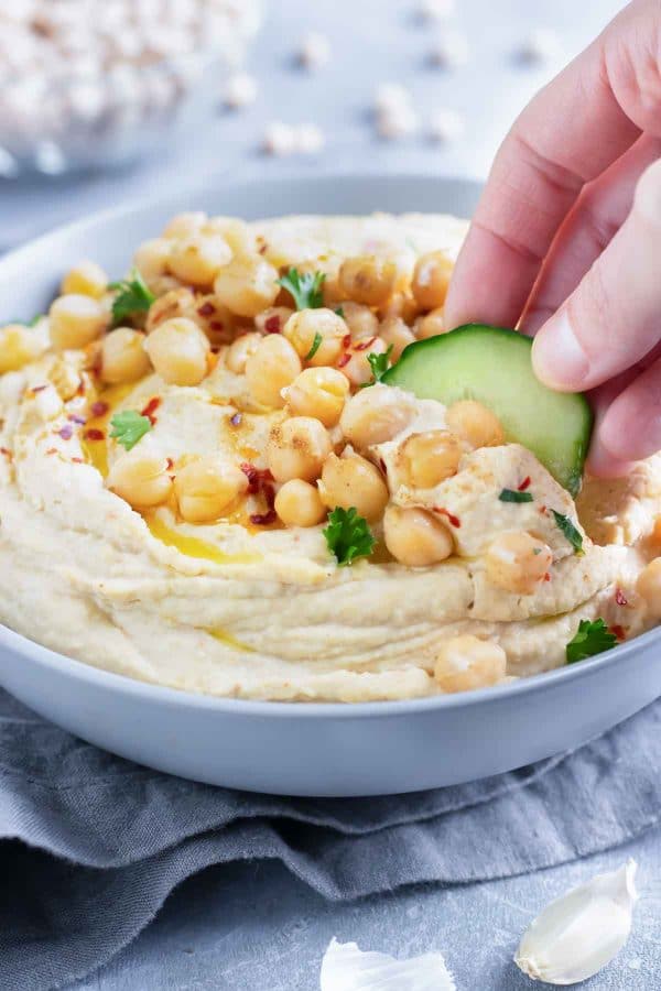 A cucumber slice being dipped into a party bowl full of a healthy authentic hummus.