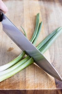 A sharp knife is used to cut the green onions on a cutting board.