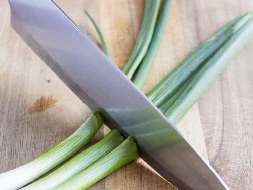 A sharp knife is used to cut the green onions on a cutting board.