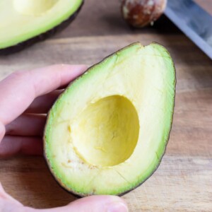 A halved avocado sits on a wooden cutting board beside a knife with a pit on it.