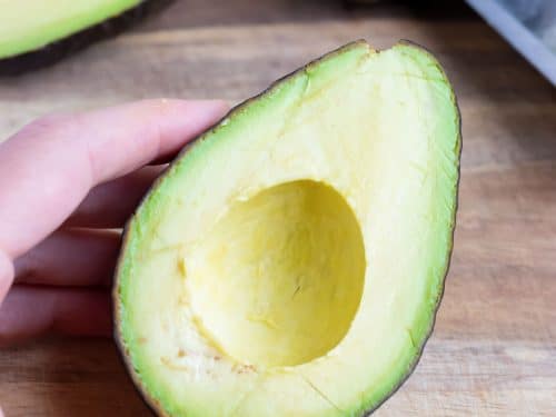 A halved avocado sits on a wooden cutting board beside a knife with a pit on it.