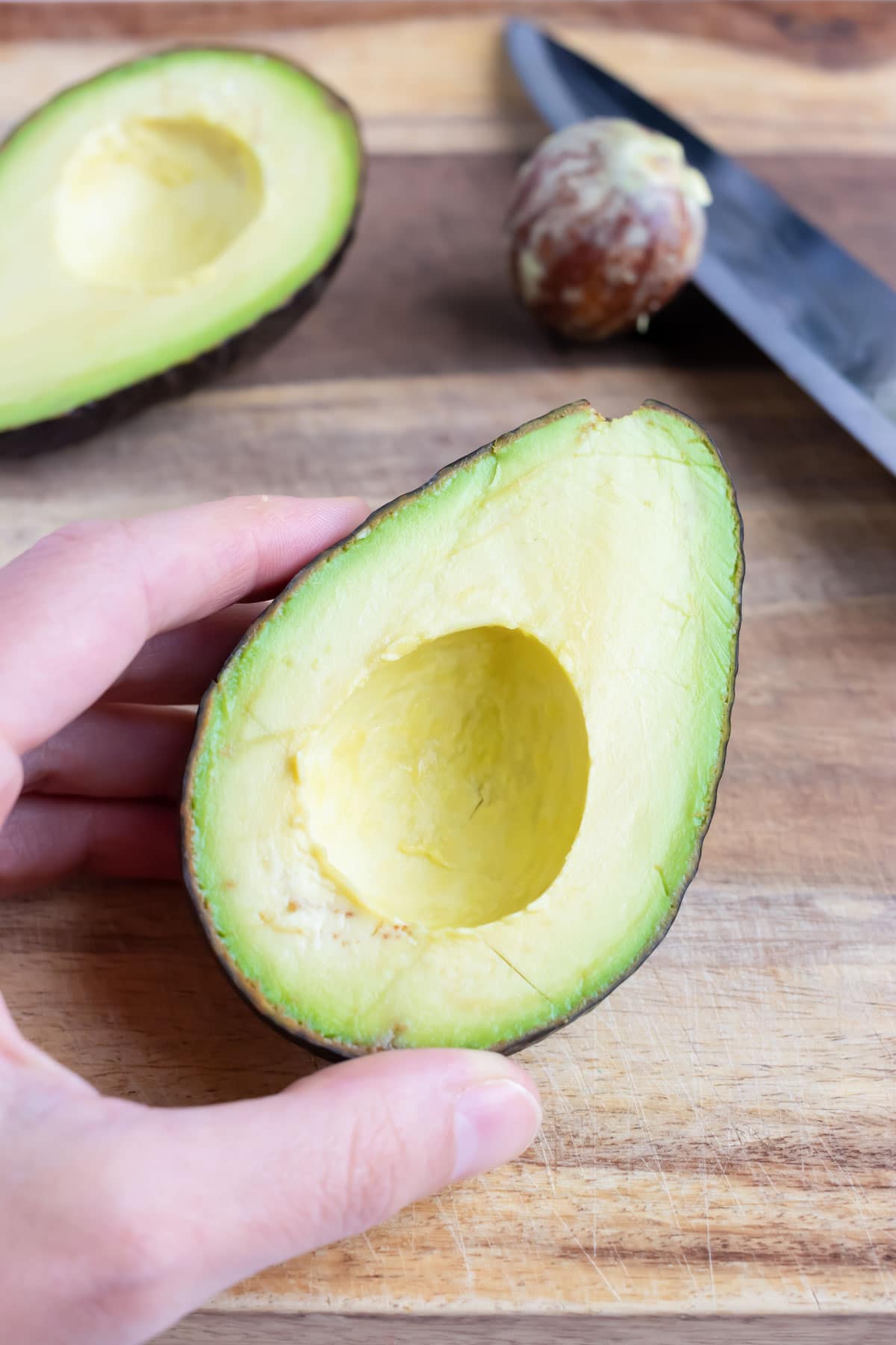 A halved avocado sits on a wooden cutting board beside a knife with a pit on it.