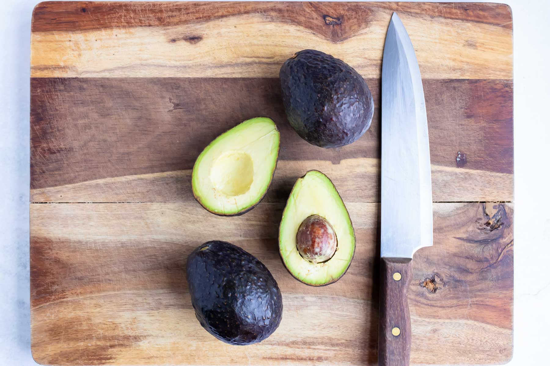 A halved avocado sits beside two more avocados and a kitchen knife on a cutting board.