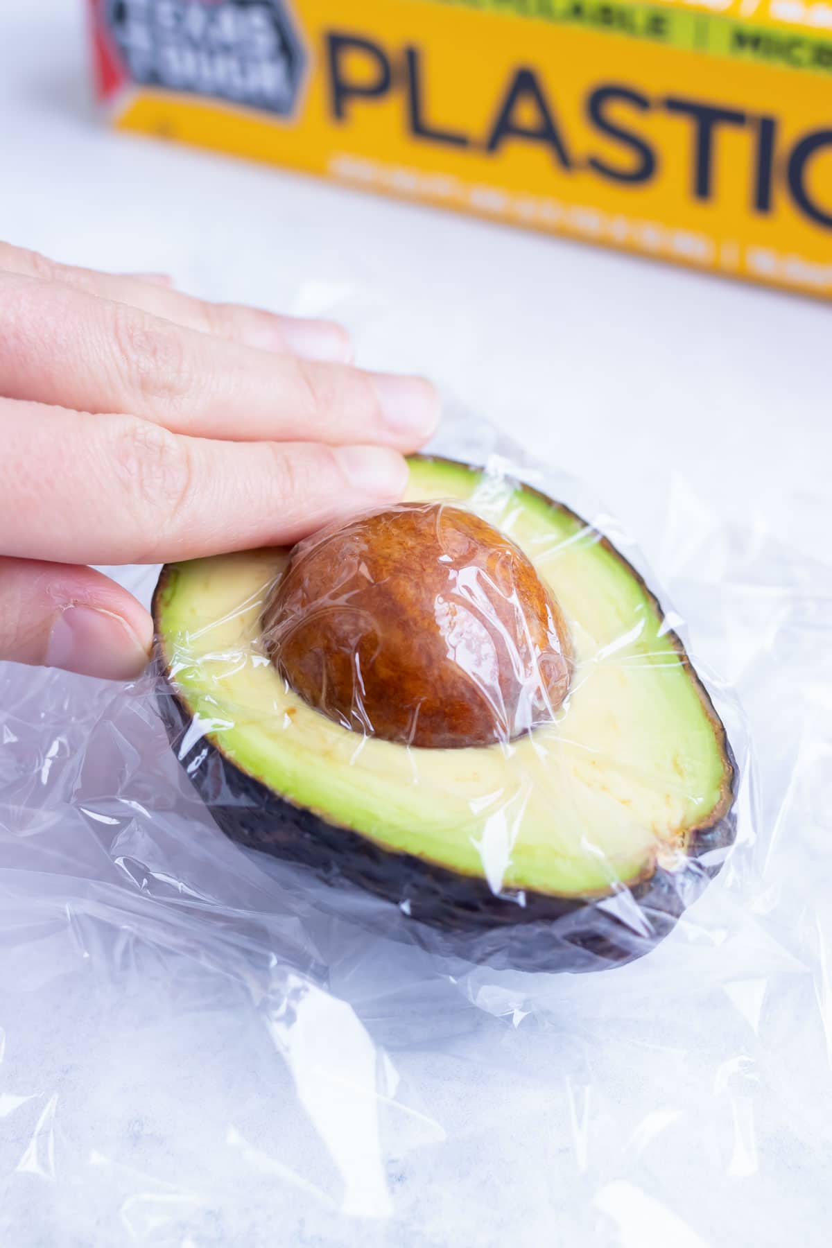 A sliced open avocado with the pit inside and a plastic ripe placed over the exposed avocado.